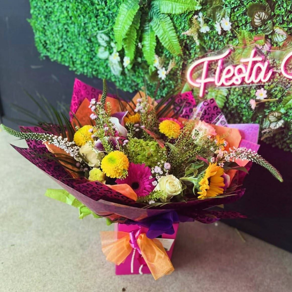 A bright bouquet in front of the shop's foliage wall and pink neon sign